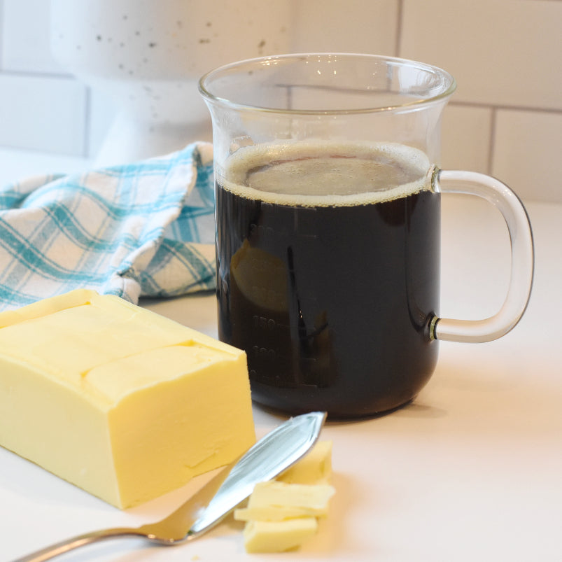 Glass mug filled with black coffee, a cube of grass-fed Kerrygold butter with a butter knife beside it with blue checkered towel in the background 