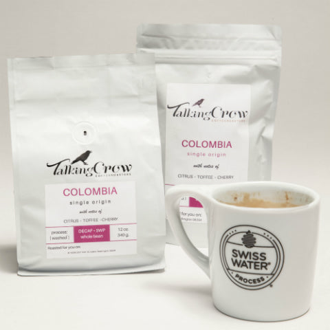 12 and 4 oz bags of Talking Crow Coffee Roasters Decaf Colombian coffee with a white Swiss Water Process mug filled with brewed coffee