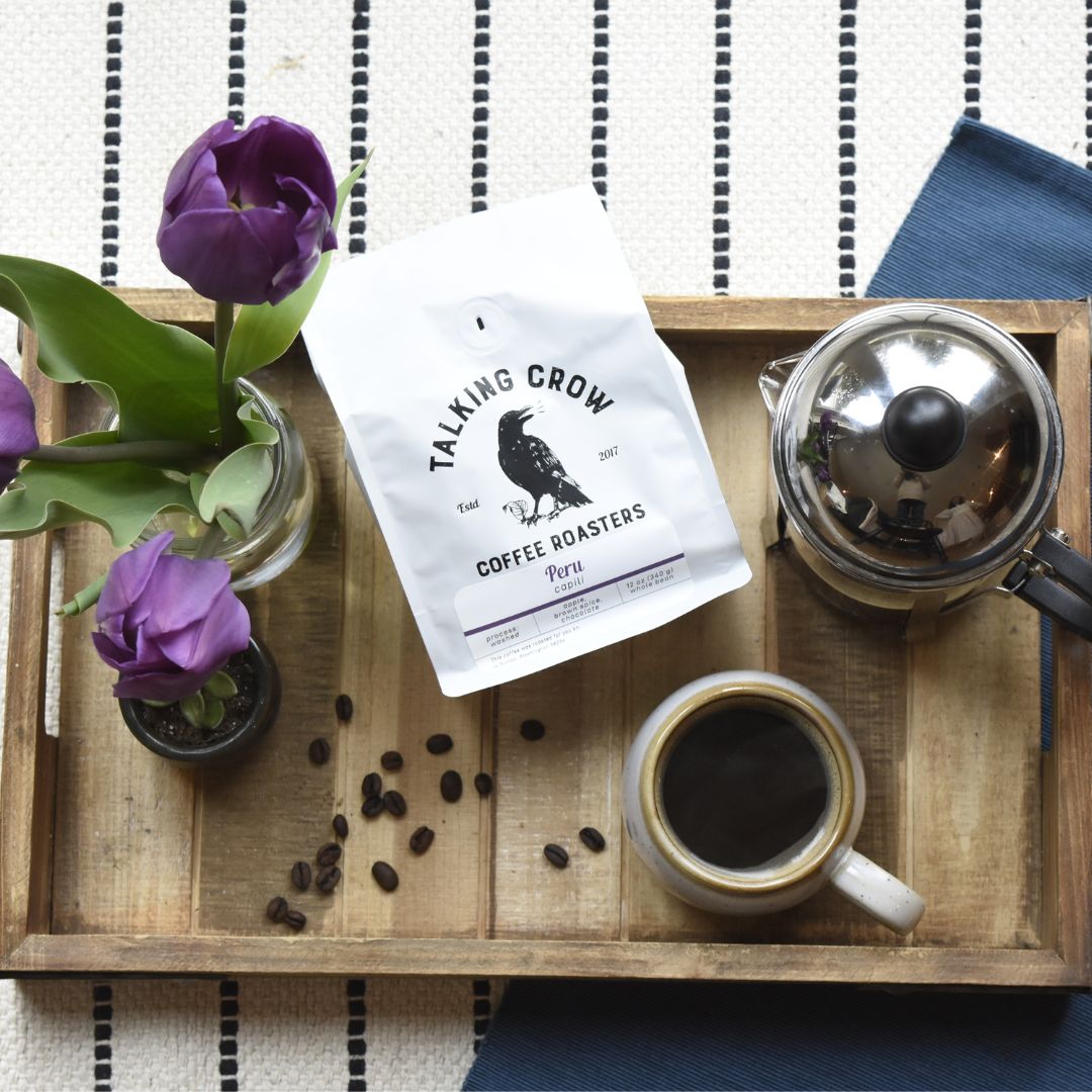Flat lay of a wooden tray with a vase of purple tulips, a 12 oz bag of Talking Crow Coffee Roasters single origin specialty decaf Peru, a mug with coffee, and a French press brewer.