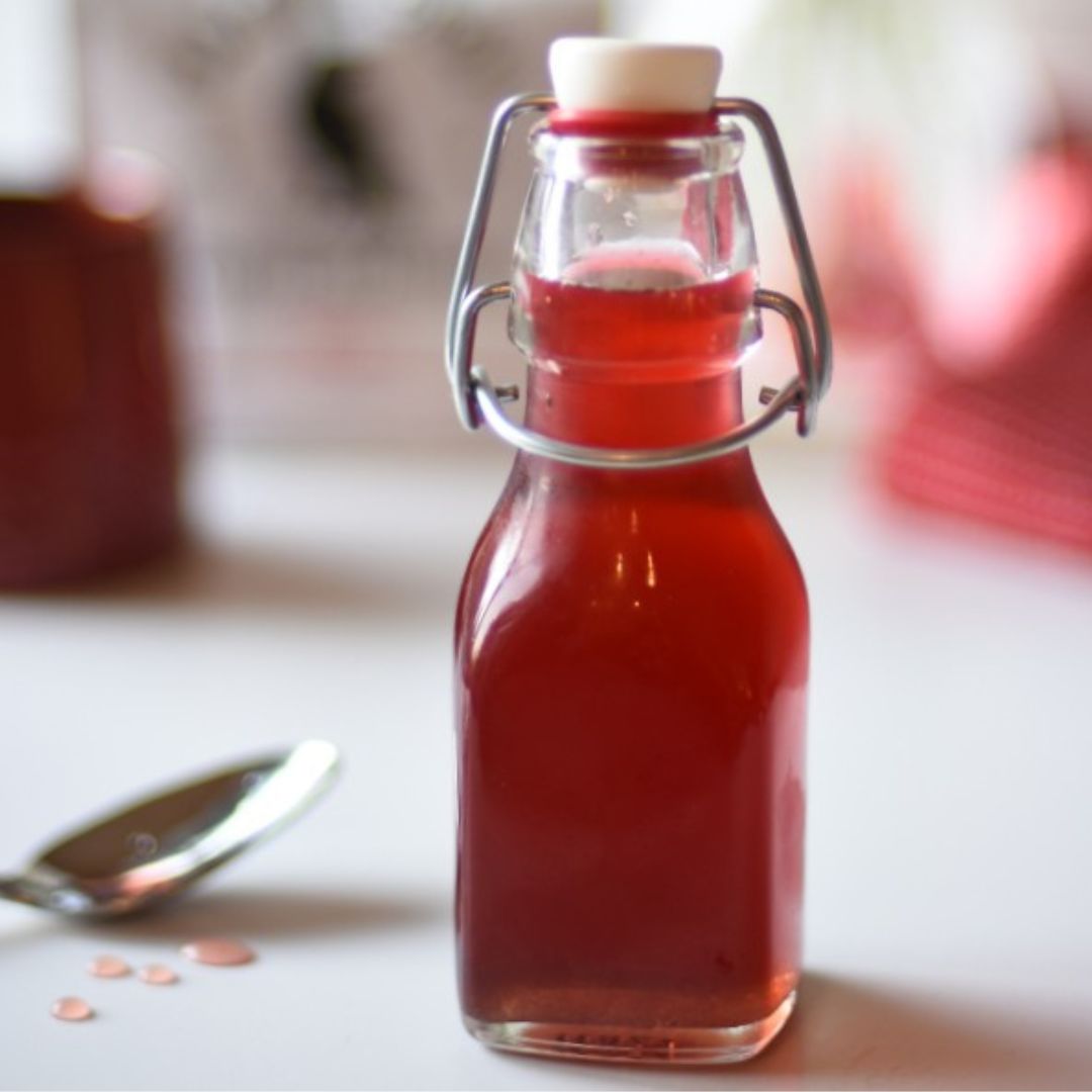 Glass flip top bottle with Rose simple syrup, a spoon with the syrup dribbled on it, and  and a bag of Talking Crow Coffee Roasters in the background.