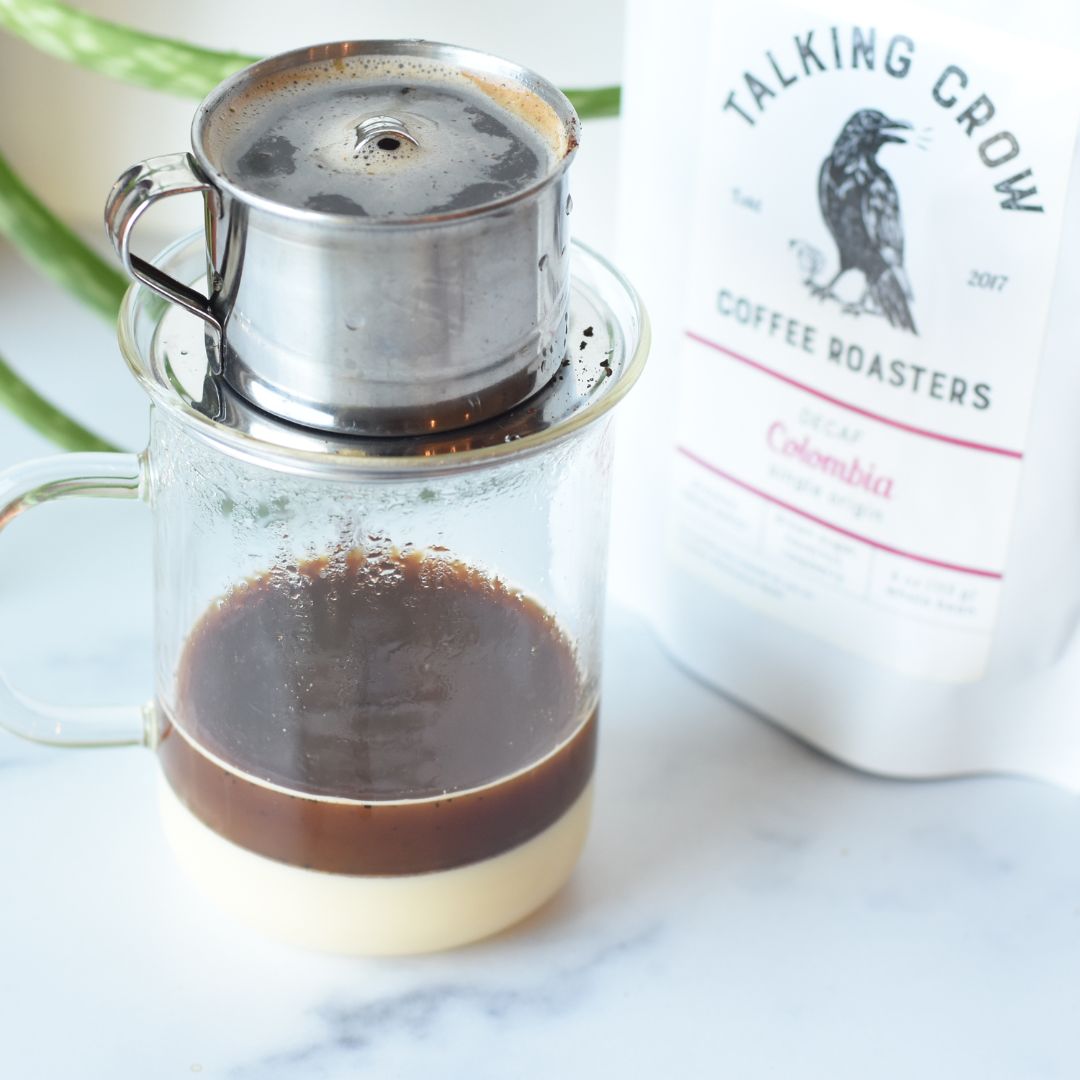 Vietnamese coffee brewer - phin on top of a glass mug with layers of sweetened condensed milk and coffee. A bag of Talking Crow Coffee Roasters single origin specialty decaf Colombia next to it.