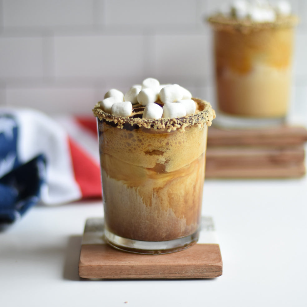 Photo of a Glass with S'mores Shaken Iced Coffee topped with mini marshmallows in the foreground, and a second blurred in the background with a red, white, and blue towel.