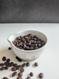 A small speckled off-white ceramic bowl filled with roasted coffee beans, with a few beans scattered around on a white surface against a dark gray background.