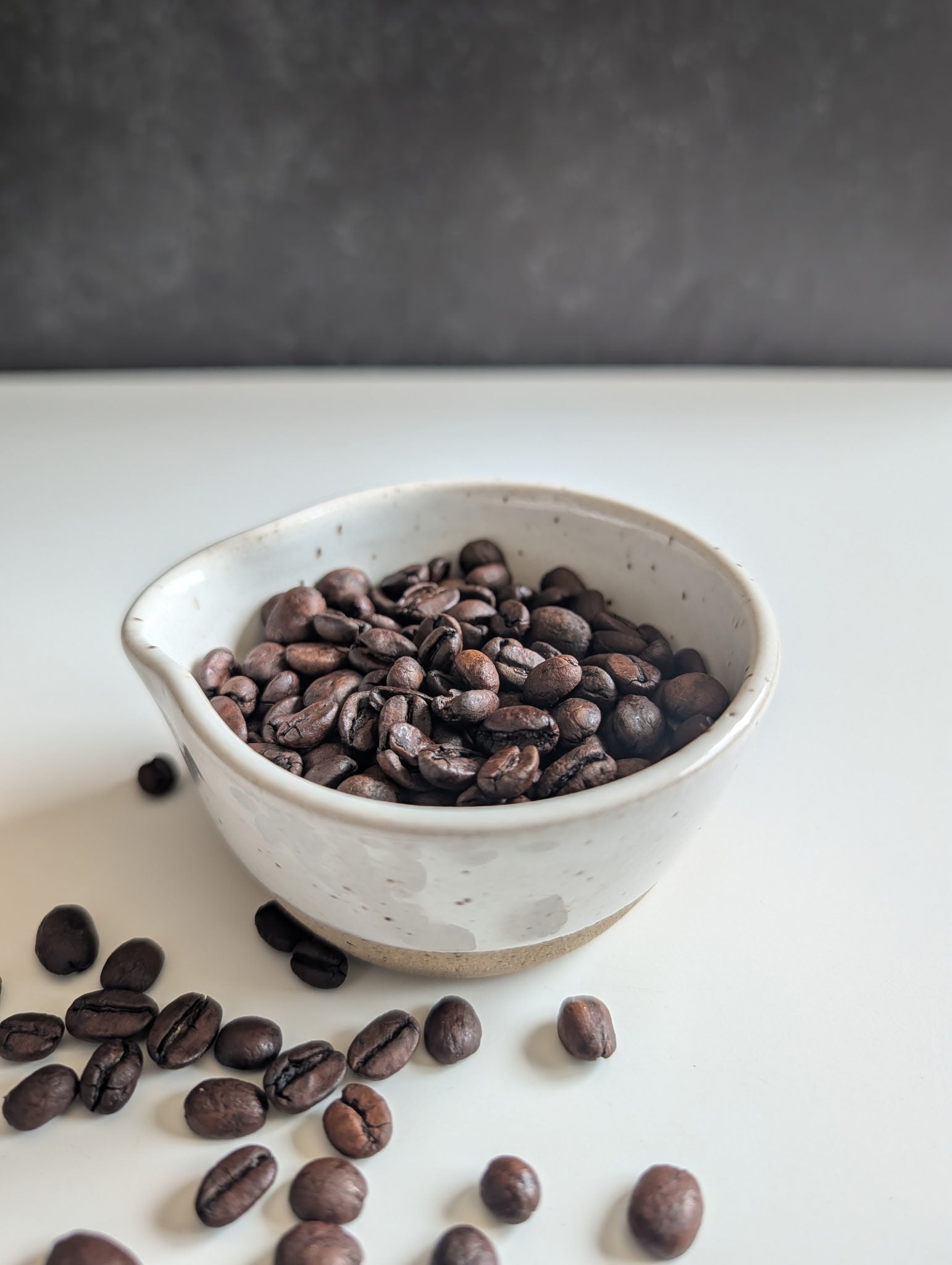 A small speckled off-white ceramic bowl filled with roasted coffee beans, with a few beans scattered around on a white surface against a dark gray background.