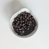 Dark roasted coffee beans filling a small white ceramic bowl, photographed from above on a clean white background.