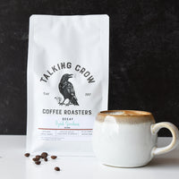 Coffee bag from Talking Crow Coffee Roasters with a mug and decaf coffee beans on a dark background